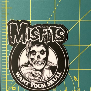 May include: Black and white sticker featuring the Misfits band logo with a skull wearing a hooded robe and the text "Want Your Skull".