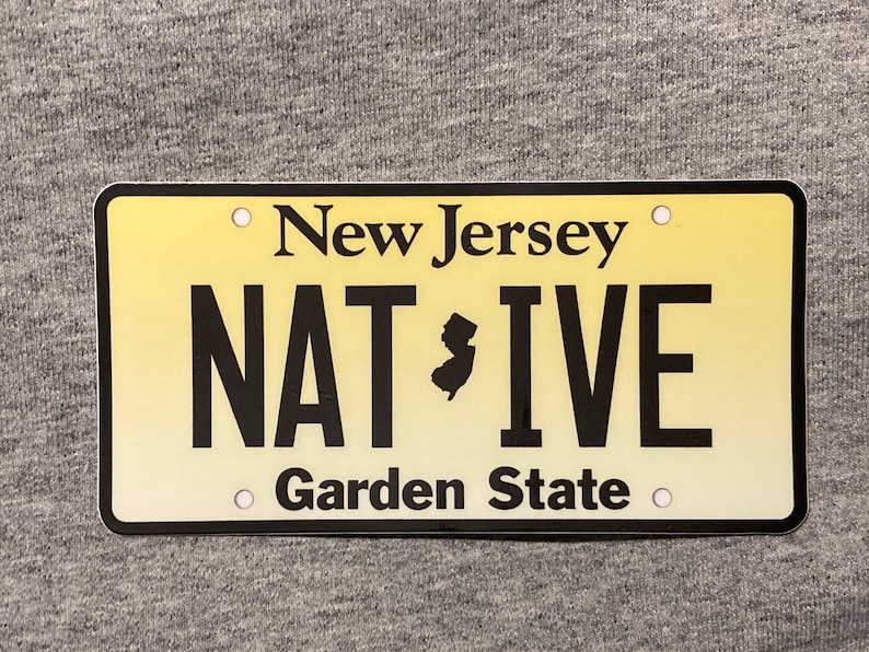 Pu&ograve; includere: Targa rettangolare con sfondo sfumato giallo. La targa riporta "New Jersey" in alto, "NAT*IVE" in grassetto nero e "Garden State" in basso. Un contorno nero incornicia la targa.