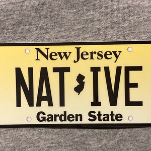 Pu&ograve; includere: Targa rettangolare con sfondo sfumato giallo. Il testo "New Jersey" &egrave; in alto, con "NATIVE" in grandi lettere nere e una silhouette del New Jersey. "Garden State" &egrave; in basso.
