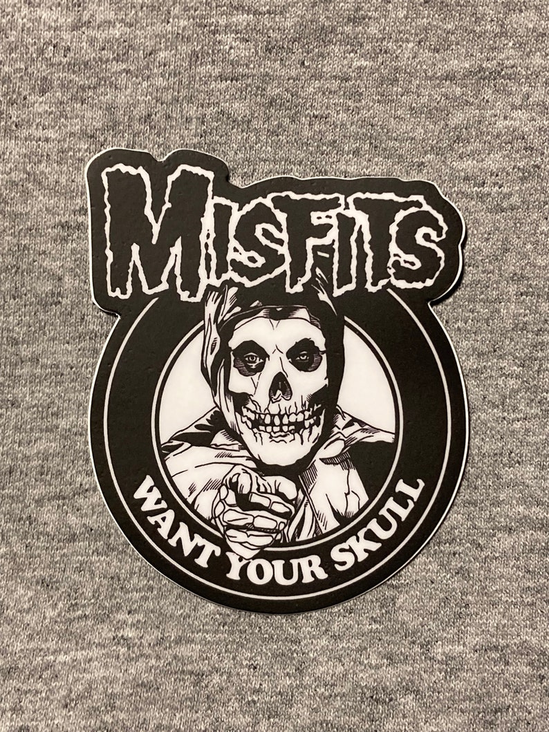 May include: Black and white sticker with the band name "Misfits" in a bold font. The sticker features a skull wearing a hooded cloak and pointing at the viewer with the text "Want Your Skull" around the image.