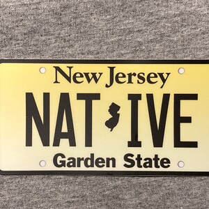 Pu&ograve; includere: Targa rettangolare con sfondo sfumato giallo. Le parole "New Jersey" sono in alto, con "NATIVE" e "Garden State" in basso. Una silhouette nera dello stato &egrave; tra "NAT" e "IVE".