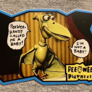 May include: A yellow puppet dinosaur with a long beak and large eyes. The puppet is wearing a red shirt and is holding a sign that says "Pee-wee's Playhouse". The dinosaur is saying "I'm not a baby!" in a speech bubble. The text "Pee-wee, Randy called me a baby!" is in a speech bubble above the dinosaur.