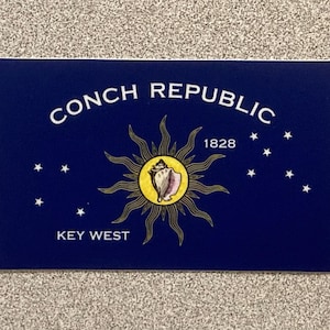 May include: A navy blue rectangular flag featuring the words "CONCH REPUBLIC" in white, arched across the top. Below, the text "KEY WEST" is centered at the bottom. The flag includes a sunburst design with a conch shell in the center and the year 1828.