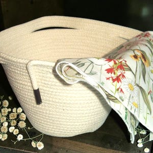 May include: A cream-colored rope basket with a handle and a floral patterned cloth. The cloth features red, yellow, and white flowers with green leaves. The basket has a dark brown accent on the side.