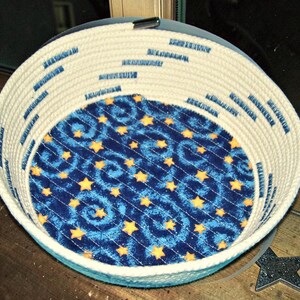 May include: A round, woven rope basket with a blue and yellow star pattern on the interior base. The basket's exterior is a cream color with blue stitching. The basket is designed for storage or decorative use.