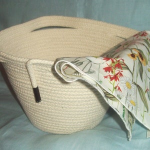 May include: A white rope basket with a floral fabric lining. The basket has two handles and a black metal accent.