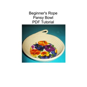 May include: A handmade rope bowl with a floral fabric base featuring orange, purple, and yellow pansies. The bowl is made of off-white rope and has a loop handle. Text on the image reads: "Beginner's Rope Pansy Bowl PDF Tutorial."