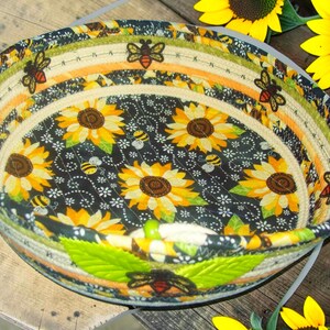 May include: A round fabric basket with a sunflower and bee pattern. The interior features a black background with yellow sunflowers and small bees. The rim has alternating stripes of green, orange, and beige, with bee and leaf accents. The basket is on a wooden surface.