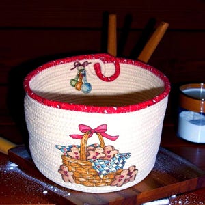 May include: A round, off-white rope basket with a red trim and handle. The basket features a painted design of a basket filled with gingerbread men and a red bow. Hanging from the handle are painted measuring spoons.
