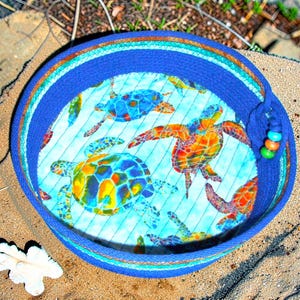 May include: A round fabric basket with a deep blue coiled rope exterior and a light blue interior lining featuring a colorful sea turtle pattern. The basket is accented with a small blue rope loop decorated with three round beads in orange, teal, and green.