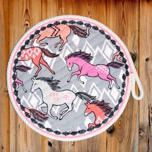 May include: Round pot holder featuring a gray background with a diamond pattern and running horses in shades of pink, coral, and white. The pot holder has a pink and black border and a white loop for hanging.