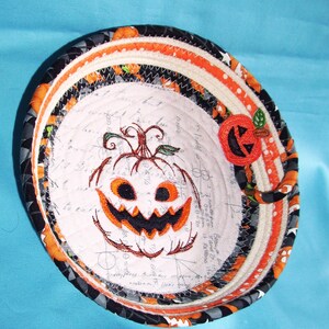 May include: A round, fabric bowl with a Halloween theme. The bowl features an embroidered jack-o'-lantern in the center, surrounded by orange, black, and white patterned fabric. A small, appliqued pumpkin decorates the rim. The bowl is approximately 6 inches in diameter.