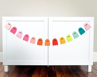 May include: A colorful ghost garland with various shades of pink, orange, yellow, green, and teal. The garland is strung across a white cabinet, creating a festive and playful decoration. Each ghost has two small black dot eyes.