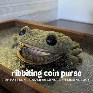 Può includere: Un portamonete a forma di rana verde all'uncinetto, con grandi occhi neri e una chiusura in metallo. Il portamonete è aperto, rivelando l'interno. Il testo "ribbiting coin purse" è visualizzato sotto la rana.