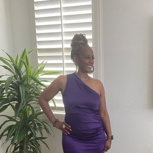 May include: A woman wearing a one-shoulder purple satin midi dress and silver heels. She is standing in front of a white wall with a window behind her.