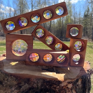 May include: A collection of wooden blocks and planks with iridescent, faceted glass inserts. The wooden pieces are arranged in a geometric display, showcasing the colorful, light-refracting properties of the glass. The wood has a warm, reddish-brown tone.