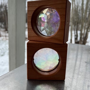 May include: Two wooden blocks stacked on top of each other. Each block has a circular opening with a kaleidoscope inside. The kaleidoscope in the top block is purple and the kaleidoscope in the bottom block is iridescent.
