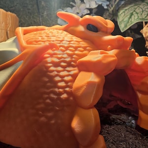 May include: Orange dragon-shaped reptile hide. The hide features a textured body, a detailed head with black eyes, and a curved archway. The dragon is a vibrant orange color, designed to provide shelter for reptiles.