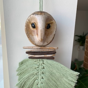 Könnte beinhalten: Ein Keramik-Eulenkopf mit brauner und weißer gesprenkelter Farbe und schwarzen Augen hängt an einer grünen Schnur. Der Eulenkopf ist an einer Reihe von braunen und weißen Holzstäben befestigt. Ein grüner Makramee-Quaste hängt an den Stäben.