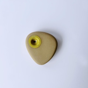 May include: A tan-colored ceramic climbing hold with a yellow center hole. The hold is shaped like a teardrop.