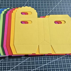Könnte beinhalten: Ein Stapel bunter, gestanzter Papiergeschenktüten in verschiedenen Farben, darunter Gelb, Rosa, Rot, Grün, Blau und Weiß. Die Tüten haben Griffe und gestrichelte Linien zum Falten.