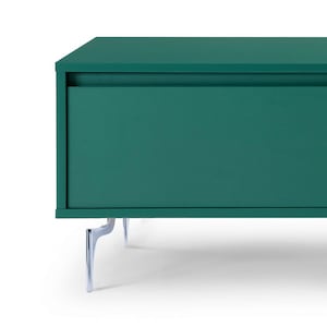 以下が含まれることがあります： 引き出しが1つとクロムメッキの脚が付いたティールグリーンのメディアコンソール。コンソールは木でできており、モダンなデザインです。
