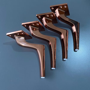 May include: Set of five copper-colored metal furniture legs with a curved design. Each leg has a square base and a white plastic foot pad.