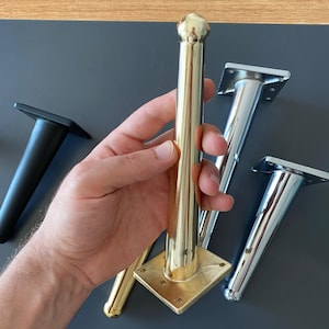 May include: Four furniture legs in different finishes: gold, silver, and black. The legs are made of metal and have a round top. The gold leg is being held in a hand.