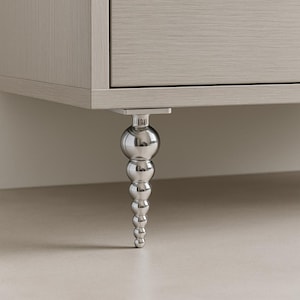 Puede incluir: Primer plano de un armario de madera de color claro con una pata decorativa plateada. La pata está hecha de múltiples esferas apiladas y está unida al armario con una placa metálica cuadrada.