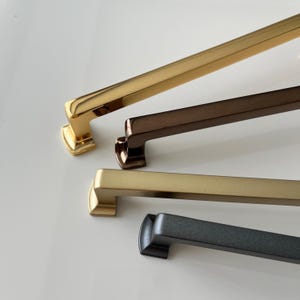 May include: Four cabinet pulls in different finishes: gold, bronze, brushed nickel, and black. The pulls are all the same style, with a rectangular shape and a slight curve at the top.