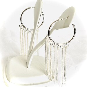 May include: Silver hoop earrings with dangling chain fringe. The earrings are displayed on a white stand against a white background. The hoops are circular and the chains hang down, creating a layered effect.