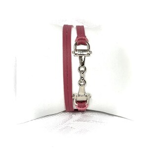 May include: A double-wrap leather bracelet with a silver-toned horse bit clasp. The bracelet is a deep pink color.