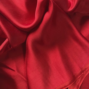 May include: A close-up of a deep red fabric with a smooth, silky texture. The fabric is folded and draped, creating soft folds and curves.