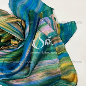 Pode incluir: Um lenço de seda com um padrão abstrato vibrante em tons de azul, verde e amarelo. O lenço tem uma aparência fluida e drapeada, com o texto "www.villagesilk.com" repetido no tecido. As palavras "Silk Vietnamsilk Handmade" também são visíveis.