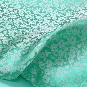 May include: A close-up of a light green fabric with a white floral pattern. The fabric is smooth and silky.
