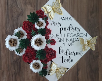 May include: A white graduation cap decorated with red, white, and gold paper flowers and gold butterfly accents. The cap has text in Spanish that reads "Para mis padres que llegaron sin nada y me lo dieron todo."