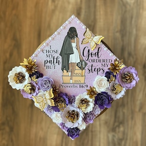 May include: A graduation cap decorated with paper flowers in gold, white, and purple. The cap features a graphic of a woman and the text "I chose my path but God ordered my steps Proverbs 16:9". Gold butterfly accents complete the design.