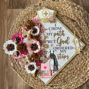 May include: A decorated graduation cap with the text "I chose my path but God ordered my steps." The cap features a nurse's hat, a syringe, and a figure of a nurse. Embellishments include pink, white, and brown paper flowers, gold butterflies, and beads.