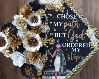 May include: Black graduation cap decorated with gold, white, and black paper flowers, pearls, and rhinestones. The cap features the text "I chose my path but God ordered my steps" and a nurse's hat graphic. The image also includes a graphic of a nurse.