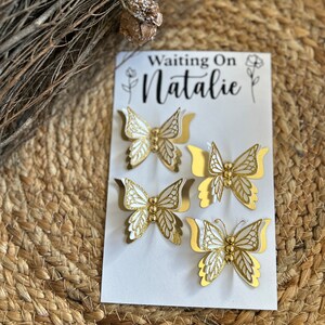 May include: A white card with the text "Waiting On Natalie" and four gold and white butterfly decorations. The butterflies have intricate cut-out details and are arranged on a woven background.