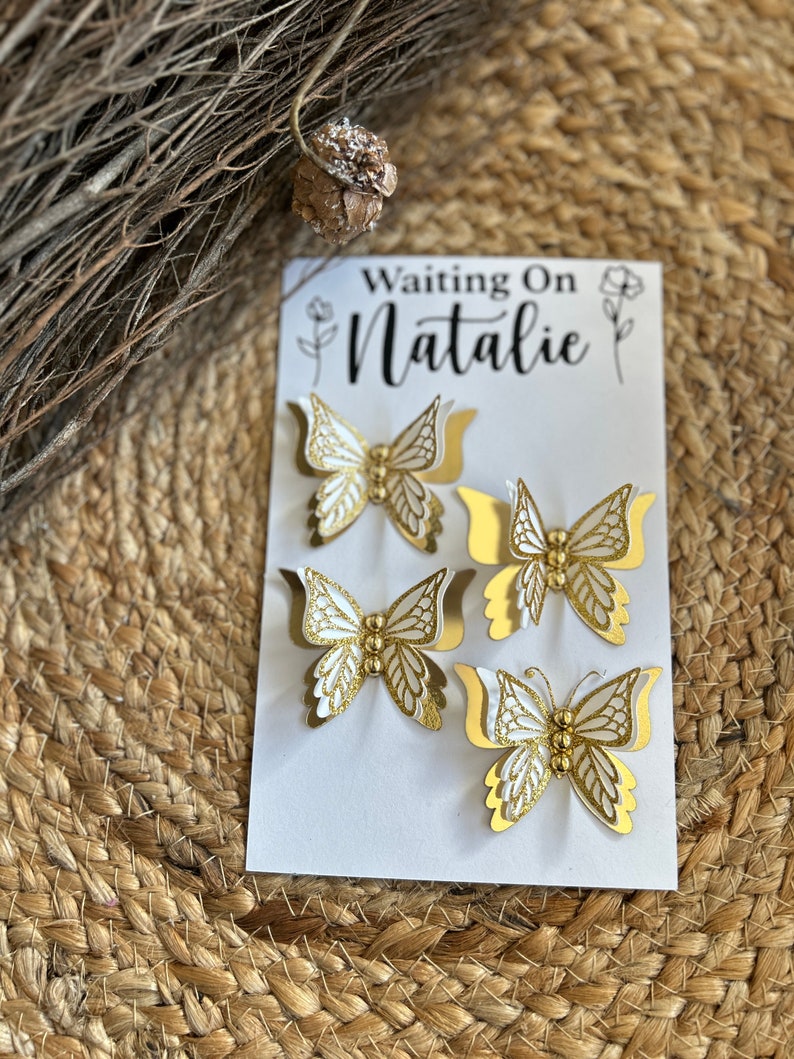 May include: A white card with the text "Waiting On Natalie" and four gold and white butterfly decorations. The butterflies have intricate cut-out details and are arranged on a woven background.