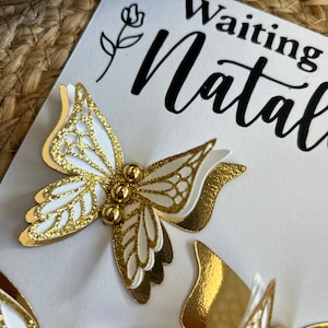 May include: A white card with gold and white butterfly embellishments. The butterflies have intricate cut-out designs and are adorned with small gold beads. The card also features the text "Waiting Natalie" in black script.
