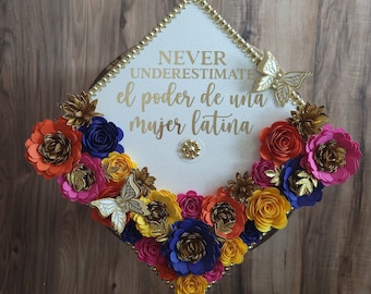 May include: A graduation cap decorated with colorful paper flowers in shades of orange, pink, yellow, and blue. The cap has gold trim and features the text "NEVER UNDERESTIMATE el poder de una mujer latina" in gold lettering, with gold butterfly accents.