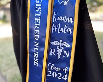May include: A blue and gold graduation sash with the words "REGISTERED NURSE" in white. The sash also features the name "Kianna Malm", a heart-shaped stethoscope, the letters "RN", and "Class of 2024".