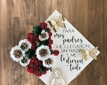 May include: A white graduation cap decorated with red, white, and gold paper flowers and two gold butterflies. The cap has text in Spanish and a gold illustration of the Virgin Mary.
