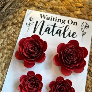 May include: A white card with the text "Waiting On Natalie" and several red paper flowers. The flowers are arranged in a cluster, with a detailed, layered design. The background is a woven, natural fiber.