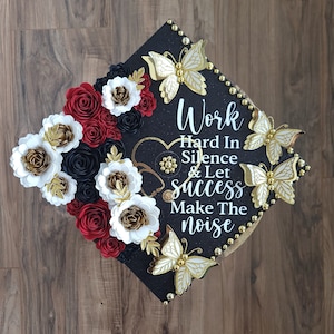 May include: Black graduation cap decorated with gold butterflies, gold beads, and paper flowers in red, white, black, and gold. The cap has the text "Work Hard In Silence & Let Success Make The Noise."