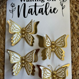 May include: Four white and gold butterfly decorations with intricate wing details and gold bead bodies are arranged on a white card. The card has the text "Waiting On Natalie" in black script, with floral accents.