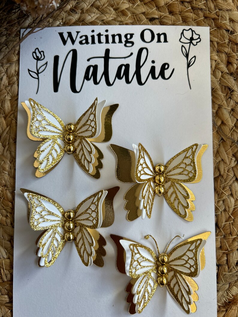 May include: Four white and gold butterfly decorations with intricate wing details and gold bead bodies are arranged on a white card. The card has the text "Waiting On Natalie" in black script, with floral accents.