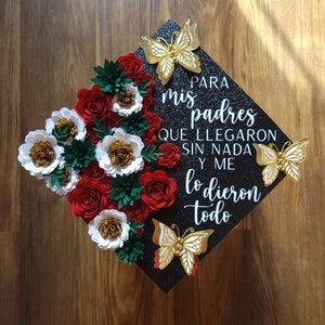 May include: Black graduation cap decorated with red, white, and gold paper flowers and gold butterflies. The cap has the Spanish text "PARA mis padres QUE LLEGARON SIN NADA Y ME lo dieron todo."